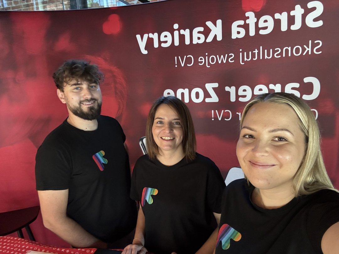 **Tekst alternatywny (PL):** Zdjęcie przedstawia trzy osoby stojące obok siebie i uśmiechające się do aparatu podczas wydarzenia promocyjnego. Wszystkie mają na sobie czarne koszulki z kolorowym symbolem w kształcie strzałki lub litery „Ł”. W tle znajduje się duży czerwony baner z częściowo widocznymi napisami dotyczącymi strefy kariery i wsparcia zawodowego. Zdjęcie zostało wykonane we wnętrzu budynku i ma nieformalny, zespołowy charakter.