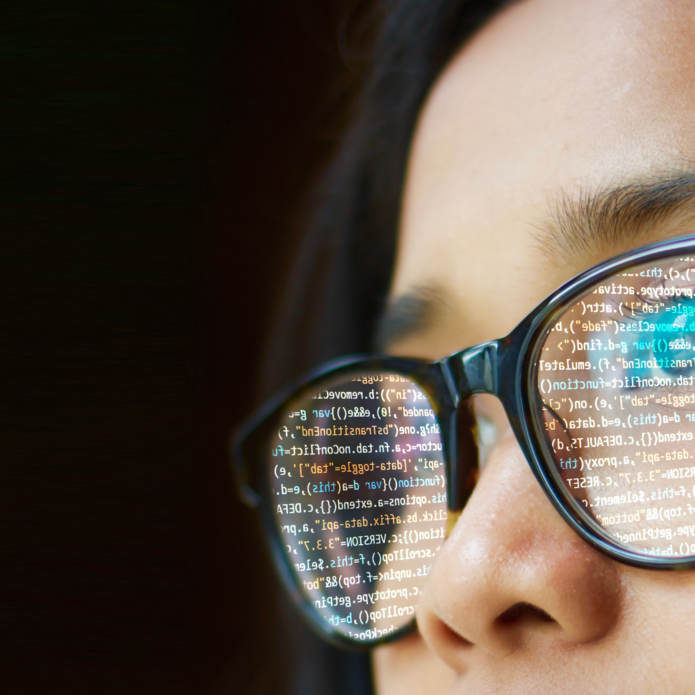 widoczna z bliska twarz i okulary w których odbija się ekran komputera z programem do kodowania